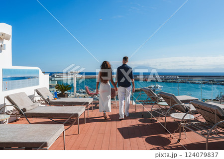A couple walks hand in hand on a terrace with lounge chairs, overlooking a marina with yachts. Clear blue sky and distant mountains suggest a coastal setting. 124079837
