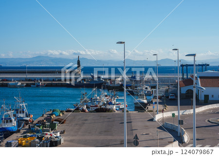 Numerous fishing boats dock along a pier in a sunny coastal town. A lighthouse stands on the breakwater, with mountains visible on the horizon. 124079867