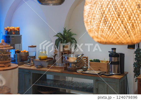 A luxury hotel interior features a wooden buffet with breakfast items, including a juice dispenser, juicer, and oranges. A woven pendant light adds warmth. 124079896