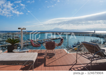 A luxurious terrace with modern furniture and a telescope overlooks a bustling marina filled with boats and yachts, set against a mountainous landscape. 124079904