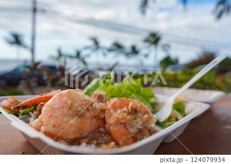 A plate of garlic shrimp over rice with greens on a wooden table, set against palm trees, ocean views, parked cars, and a blue sky in Hawaii. 124079934