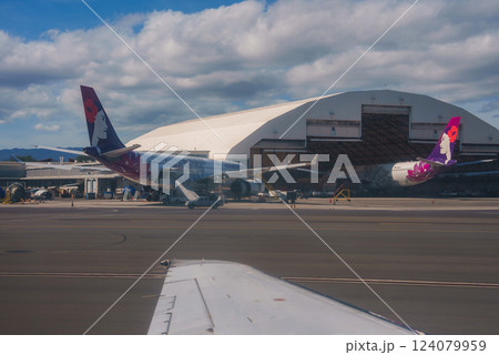 View of two Hawaiian Airlines planes parked near a white hangar, with mountains in the background and a partly cloudy sky, seen from another aircraft. View of two Hawaiian Airlines planes parked near a white hangar, with mountains in the background and a partly cloudy sky, seen from another aircraft. 124079959