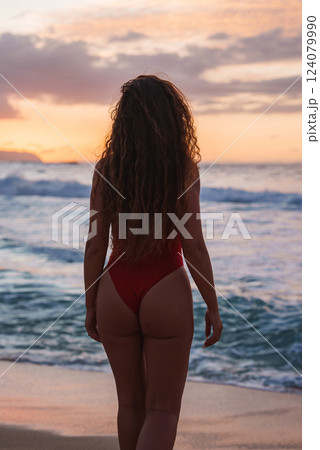 A woman with curly hair stands on a sandy beach in a red swimsuit, facing the ocean at sunset. Warm orange and pink hues fill the sky, with land visible on the horizon. 124079990