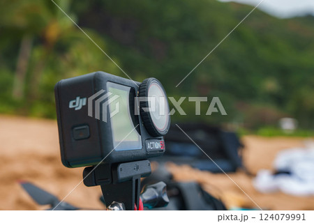 A DJI Action 5 camera is mounted on a tripod with a blurred background of sandy beach and lush green vegetation, suggesting a tropical Hawaii setting. 124079991