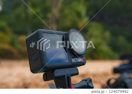 A DJI Action 3 camera mounted on a stabilizing grip, set against a blurred background of lush greenery and sandy terrain, suggesting a tropical location. 124079992