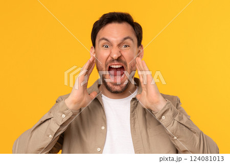 A man with brown hair and a beard is shouting with his mouth open and his hands cupped. He is wearing light brown button-down shirt over white t-shirt. He is standing in front of yellow background. 124081013