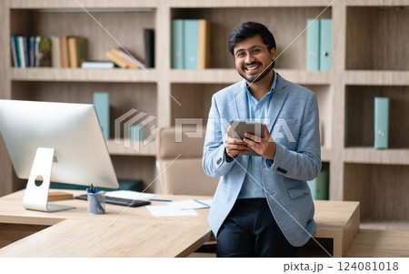 A smiling Indian businessman wearing a blue blazer stands in a modern office, leaning against a desk with a computer monitor in the background. He is holding a tablet in his hands 124081018