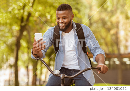 Cheerful African American Guy Having Video Call While Driving Bicycle Outdoors, Holding Smartphone And Smiling, Selective Focus 124081022