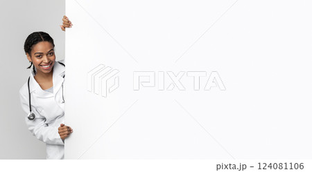 Young black female medical intern looking out behing white advertising board with free space for text and smiling, standing over light background Young black female medical intern looking out behing white advertising board with free space for text and smiling, standing over light background 124081106