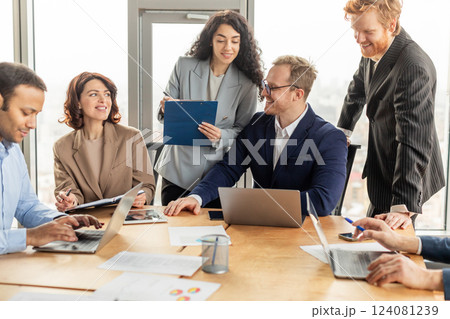 A diverse group of business professionals gathered around a table in a modern office, collaborating on a project. Two people are focused on their laptops, while another holds a clipboard with papers 124081239