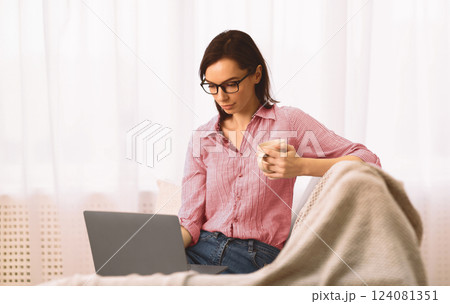Telecommuting Concept. Focused Girl In Glasses Sitting On Couch And Working On Laptop, Drinking Tea, Copyspace Telecommuting Concept. Focused Girl In Glasses Sitting On Couch And Working On Laptop, Drinking Tea, Copyspace 124081351