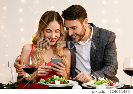 Young Couple Using Smartphone Together During Romantic Dinner In Restaurant, Browsing Internet, Social Networks Or Watching Photos. Free Space Young Couple Using Smartphone Together During Romantic Dinner In Restaurant, Browsing Internet, Social Networks Or Watching Photos. Free Space 124081480