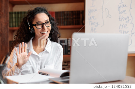 Distance Education. Positive lady wearing glasses and wireless headest at virtual meeting, sitting at desk, having video call on laptop, waving to webcam. Woman studying or teaching online at home 124081552