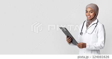 Smiling black muslim female doctor in hijab holding clipboard with clinical story of patient, posing over light background with free space 124081626