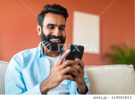 Portrait Of Cheerful Indian Man Using Mobile Phone Surfing Internet, Networking And Texting Sitting On Sofa Indoors. Young Bearded Guy Messaging And Communicating Via Smartphone Gadget Online 124081653