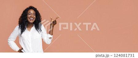 A young African American woman with long curly hair smiles while pointing to the right. She is wearing a white shirt and standing in front of a peach-colored background. A young African American woman with long curly hair smiles while pointing to the right. She is wearing a white shirt and standing in front of a peach-colored background. 124081731