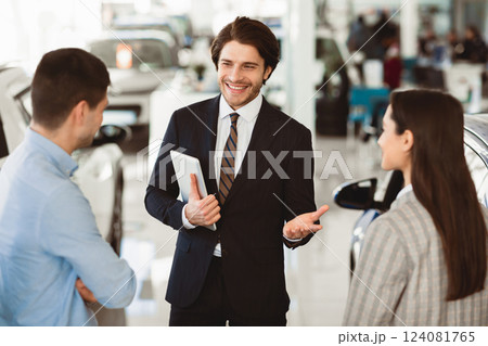 Professional Car Seller Talking With Young Couple Clients Selling Auto Working In Dealership Center. Selective Focus 124081765