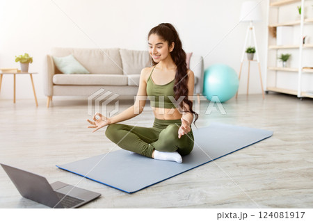 Beautiful Indian woman meditating to online video on laptop computer at home. Young Eastern lady doing yoga to reach inner balance and peace, sitting in lotus pose, performing breathing exercises 124081917