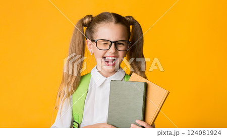 Funny Elementary School Girl Winking At Camera Holding Books Posing Over Yellow Background In Studio. Happy Back-To-School Funny Elementary School Girl Winking At Camera Holding Books Posing Over Yellow Background In Studio. Happy Back-To-School 124081924