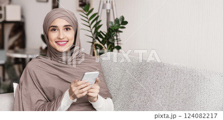 Portrait of smiling muslim woman in hijab using smartphone at home, relaxing on couch and looking at camera, free space Portrait of smiling muslim woman in hijab using smartphone at home, relaxing on couch and looking at camera, free space 124082217