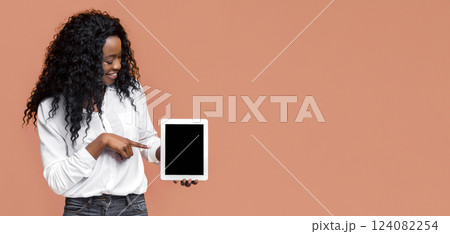 A smiling black woman with curly dark hair is wearing a white button-down shirt and dark blue jeans. She is holding a tablet in her left hand and pointing at the screen with her right index finger A smiling black woman with curly dark hair is wearing a white button-down shirt and dark blue jeans. She is holding a tablet in her left hand and pointing at the screen with her right index finger 124082254