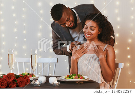 Gallant african american man putting golden necklace on his woman, having valentine dinner at restaurant, empty space Gallant african american man putting golden necklace on his woman, having valentine dinner at restaurant, empty space 124082257
