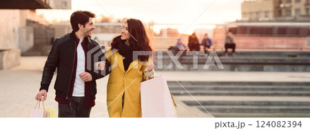 A couple walks hand in hand, smiling and carrying shopping bags as the sun sets over a city waterfront, copy space A couple walks hand in hand, smiling and carrying shopping bags as the sun sets over a city waterfront, copy space 124082394