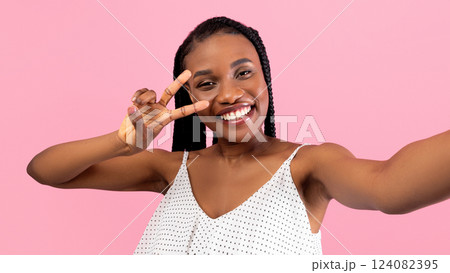 Charming young black woman showing peace gesture while taking selfie on pink studio background. Smiling African American lady making photo of herself on mobile phone Charming young black woman showing peace gesture while taking selfie on pink studio background. Smiling African American lady making photo of herself on mobile phone 124082395