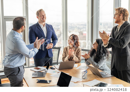 A group of four business professionals are gathered around a conference table, clapping and smiling at a colleague who is standing. The group is in a modern office with large windows 124082418