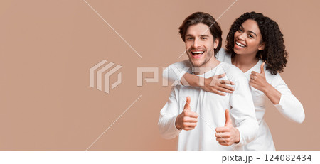 Cool. Portrait of happy interracial couple embracing and gesturing thumbs up over yellow background in studio, empty space 124082434