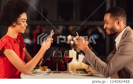 Modern Relationship. Happy Afro Couple Using Smartphones During Romantic Date In Restaurant. Panorama Modern Relationship. Happy Afro Couple Using Smartphones During Romantic Date In Restaurant. Panorama 124082488