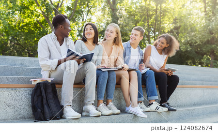 friendly multiethnic group of students discussing new educational project, sitting with notepads on stairs in park 124082674