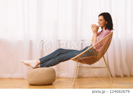 Morning Relaxation. Girl sitting in armchair putting legs on ottoman and enjoying coffee against window, copyspace Morning Relaxation. Girl sitting in armchair putting legs on ottoman and enjoying coffee against window, copyspace 124082679