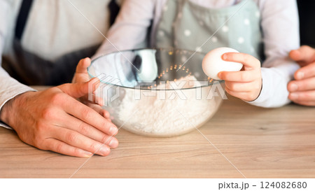 Father and daughter preparing dough together in kitchen, adding eggs to bowl while cooking pastry at home, cropped image, closeup 124082680
