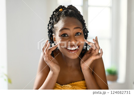 A young black woman with braided black hair smiles brightly while applying moisturizer to her face. She is standing in a bathroom with a white tiled wall and a window in the background. 124082731
