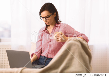 Freelance. Casual beautiful young woman in eyeglasses working on a laptop sitting on the couch at home, copyspace 124082831