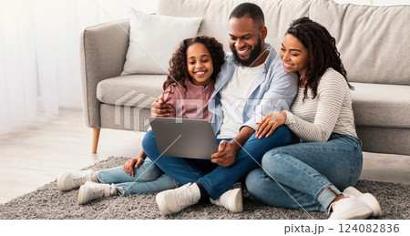 Family Weekend. Cheerful African American parents and their little daughter using laptop at home together, watching movie or browsing internet, sitting on the floor carpet in living room 124082836