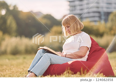 Sitting on the armchair and reading the book. Senior woman having nice weekend outdoors on the field at sunny day 124082858