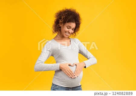 Happy expectation. Afro woman making heart shape on pregnant belly, standing over pink background 124082889