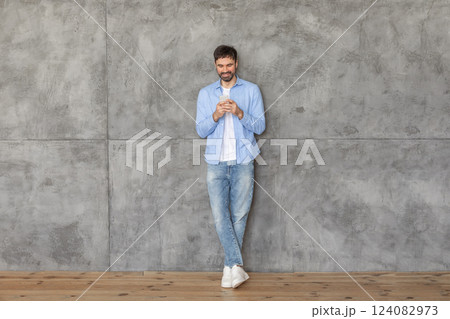 A young man in a blue shirt and jeans stands with his back against a concrete wall. He is using his smartphone and smiling as he looks down at the screen. The floor is made of wood. 124082973