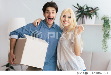 A young couple stands in their new home, smiling and holding a box and keys. The man is holding the box and the woman is holding the keys. A young couple stands in their new home, smiling and holding a box and keys. The man is holding the box and the woman is holding the keys. 124082994