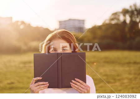 Holding book and looking into the camera. Young girl is on the field at sunny daytime having nice weekend Holding book and looking into the camera. Young girl is on the field at sunny daytime having nice weekend 124083092