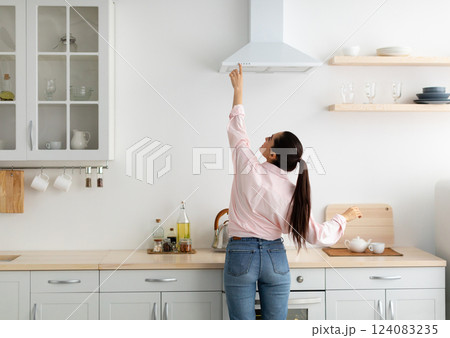 Household Concept. Back rear view of casual lady choosing and selecting mode on range exhaust hood, pushing button on mechanical fan above the stove. Female standing cooking in modern kitchen at home 124083235