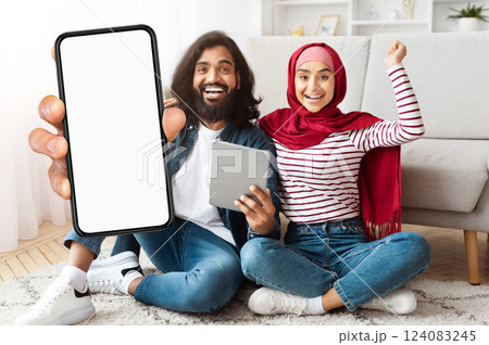 A joyful eastern couple is seated on the floor of their living room, excitedly celebrating. One person holds up a smartphone, while the other smiles and holds a tablet A joyful eastern couple is seated on the floor of their living room, excitedly celebrating. One person holds up a smartphone, while the other smiles and holds a tablet 124083245