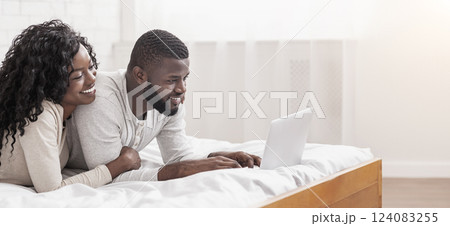 Cheerful african american spouses lying in bed using laptop together, relaxing at home. 124083255