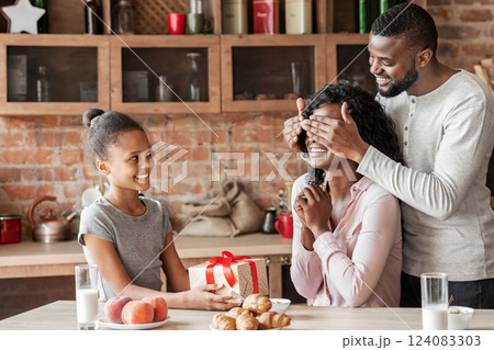 Birthday, mother day concept. Cheerful african little girl holding gift for mother, smiling man closing woman eyes, kitchen interior, free space 124083303