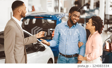 New Car. Salesman Giving Automobile Key To Happy Afro Couple Customers Standing In Auto Dealership Center. Panorama New Car. Salesman Giving Automobile Key To Happy Afro Couple Customers Standing In Auto Dealership Center. Panorama 124083334