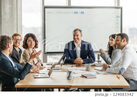 A group of business professionals sit around a large wooden conference table, engaged in a meeting. They are looking at a large screen behind them displaying a projected growth graph A group of business professionals sit around a large wooden conference table, engaged in a meeting. They are looking at a large screen behind them displaying a projected growth graph 124083342