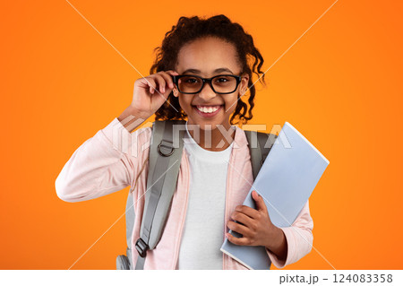 Closeup portrait of confident cheerful black female pupil wearing and touching eyeglasses, holding educational materials, posing and looking at camera isolated on yellow orange studio background wall Closeup portrait of confident cheerful black female pupil wearing and touching eyeglasses, holding educational materials, posing and looking at camera isolated on yellow orange studio background wall 124083358