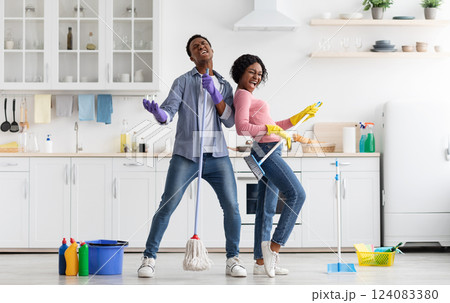 Joyful black loving couple singing songs while cleaning kitchen, using broom and mop as microphones and guitar, cheerful african american young man and woman imitating rock stars while house-keeping 124083380